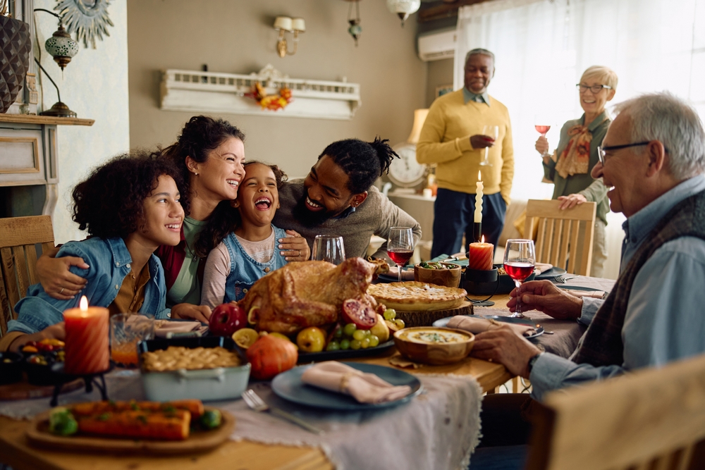 Happy multiracial parents and their kids laughing during family meal on Thanksgiving in dining room.
