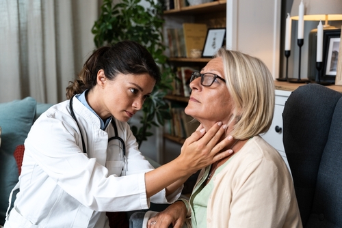 During a home visit to an elderly patient, a young endocrinologist doctor checks her thyroid gland by feeling her neck with her hands. Senior women endocrinology health care
