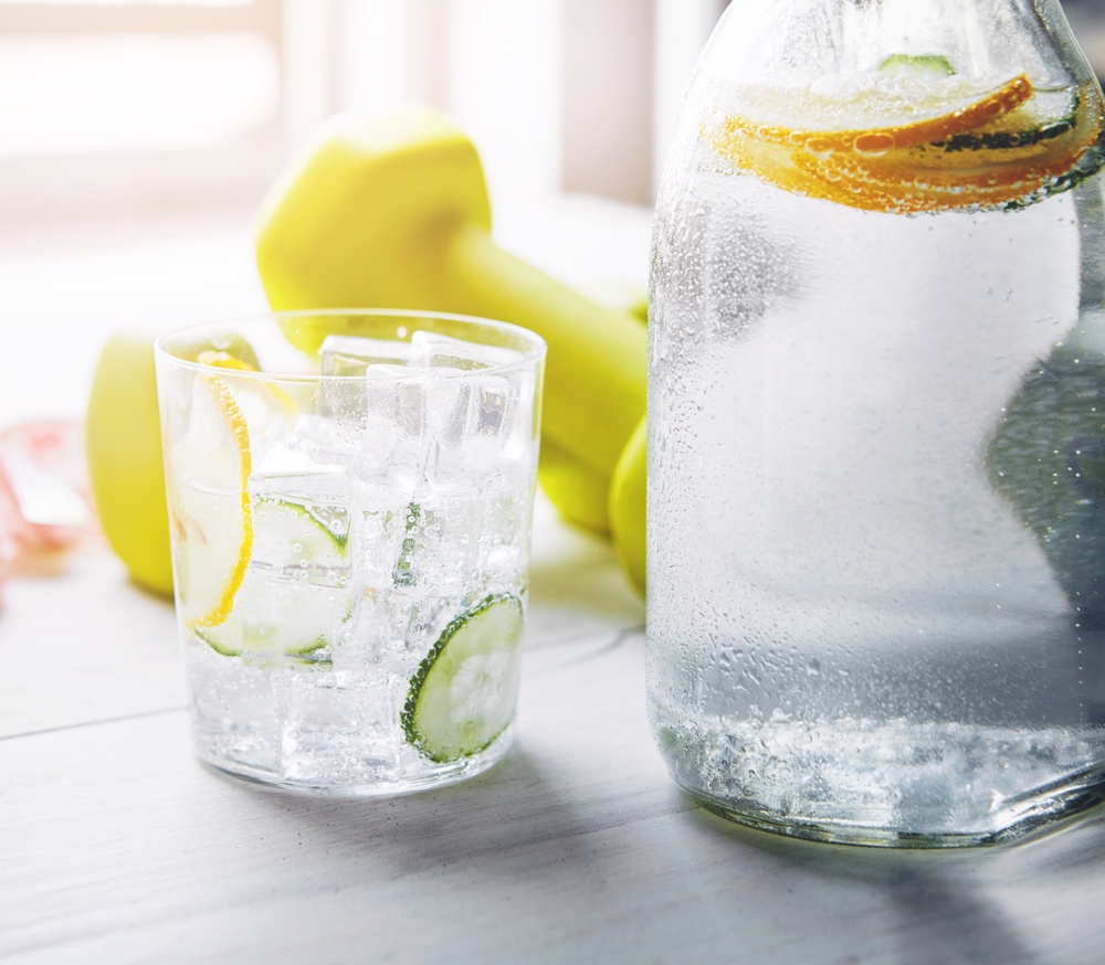 Cucumber, lemon and water in glass with dumbbell on table for vitamin C, nutrition and organic hydration. Citrus, fitness and ice with liquid in clear jug for antioxidants, detox and healthy drink