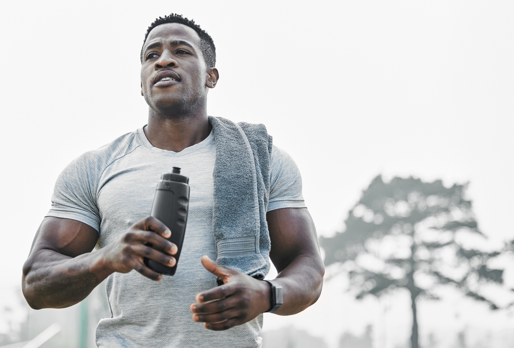 Black man, break and drinking water outdoor for fitness, wellness and body health goals in morning. Male athlete, bottle and hydration with liquid and towel on field for workout, training or exercise
