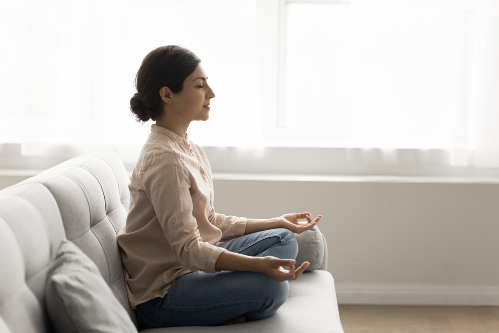 30s woman sit on sofa meditating at home, calming mind, relax body, reducing stress levels, promoting sense of inner peace, improve mental clarity, practicing mindfulness and deep breathing techniques
