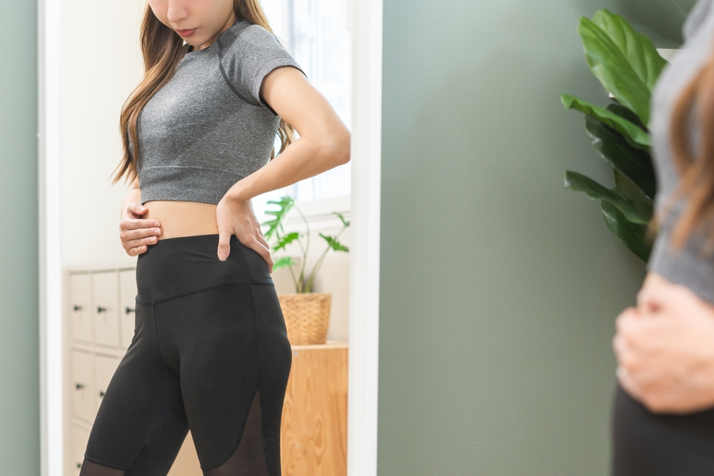 Excess weight gain, overweight asian young woman touching stomach wearing fit body clothes, dissatisfied about her body shape from fat around waist. Obese getting belly fat, diet, weight loss concept
