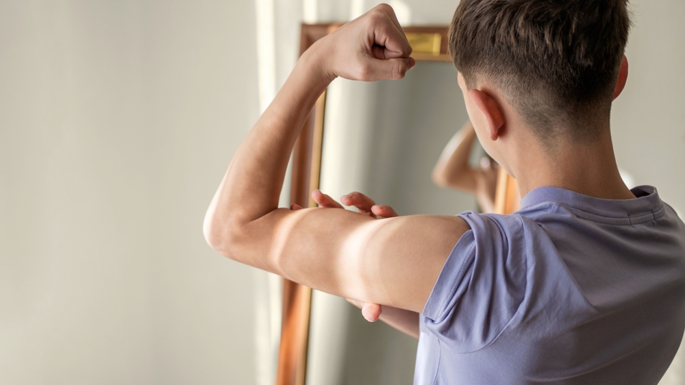 Teenage boy looks at muscle of his arm in mirror, concerned about his musculature and physique, muscle-building obsession, body dysmorphism disorder, masculinity and muscularity
