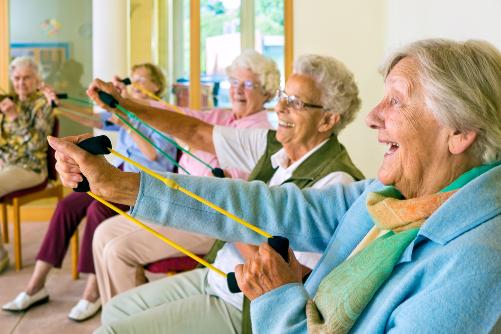 Large group of happy enthusiastic elderly ladies exercising in a gym sitting in chairs doing stretching exercises with rubber bands