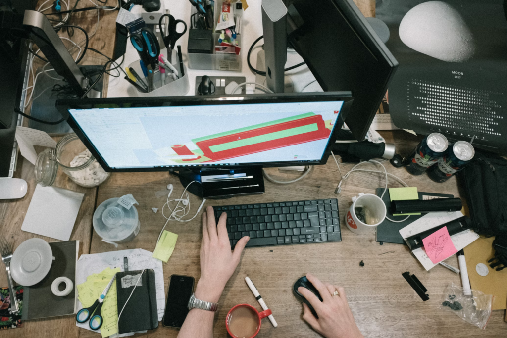 An individual focused on a computer, utilizing a mouse and keyboard, surrounded by a messy and inspiring workspace.