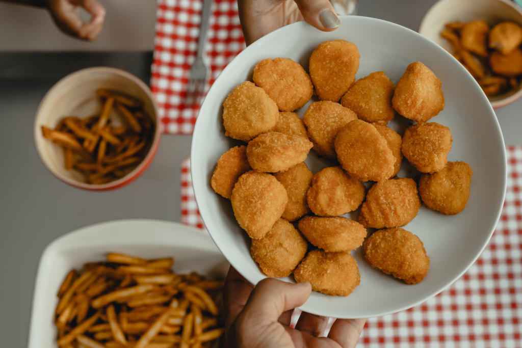 A hand presents a plate of fried chicken nuggets, emphasizing the health concerns related to advanced glycation end products.
