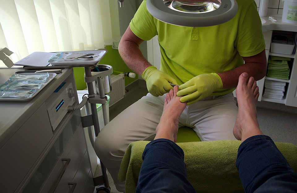 A podiatrist in a bright green shirt and gloves trims a patient's toenail under a magnifying lamp in a clinical setting, highlighting professional foot care.