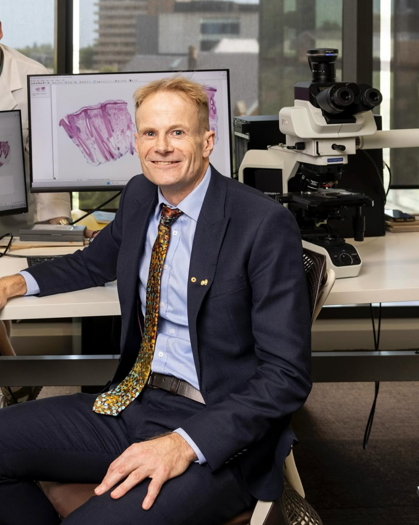 A man in a navy suit with a colorful tie sits at a desk in a laboratory, smiling at the camera. Behind him, a microscope and computer monitors display magnified tissue samples. Natural light streams through large windows showing a cityscape in the background.