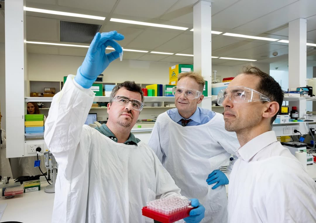 Three scientists in a laboratory wearing protective eyewear and lab coats. One man in blue gloves holds up a small vial while the other two observe closely. Laboratory equipment and supplies are visible in the background.