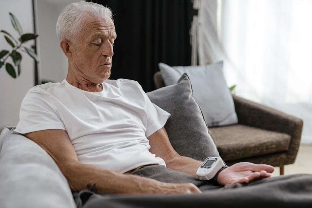 An elderly man with white hair is sitting on a couch, wearing a white t-shirt. A blood pressure monitor is attached to his left wrist, and he has a blanket draped over his lap. The room is softly lit with natural light coming through sheer curtains, and there are decorative pillows and a plant in the background, creating a calm and comfortable atmosphere.