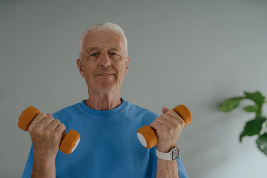 An older man with white hair is engaged in a light workout, holding two small orange dumbbells. He is wearing a blue t-shirt and a smartwatch on his left wrist. His expression is focused yet content, suggesting he is enjoying the activity. The background is simple and neutral, with a hint of greenery from a plant on the right side, adding a touch of nature to the scene.