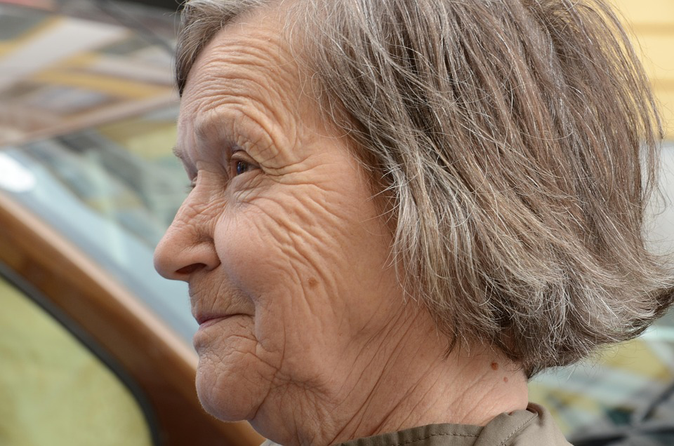 A close-up profile of an elderly woman with short gray hair and wrinkles, gazing forward with a gentle smile. A blurred car and buildings are visible in the background.