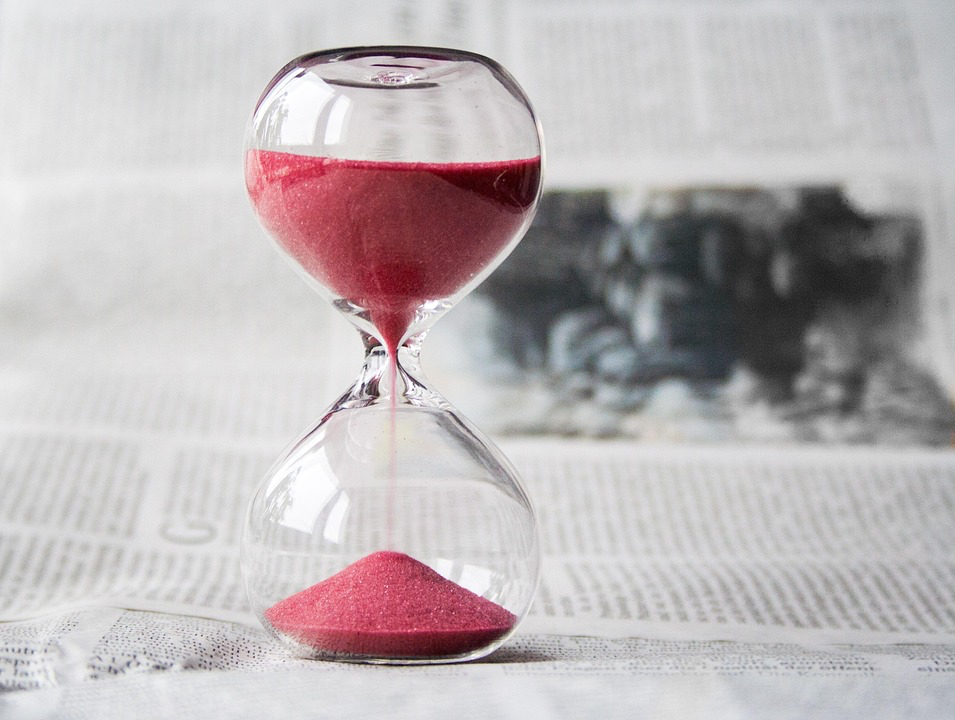 A close-up of an hourglass filled with vibrant pink sand, symbolizing the passage of time. The sand is slowly trickling from the top chamber to the bottom, creating a mesmerizing visual of time slipping away. The hourglass rests on a surface covered with text, possibly a newspaper or book, adding a layer of intellectual depth to the scene. This image evokes a sense of urgency and the fleeting nature of moments, much like watching a Pistachio Tree grow over time.