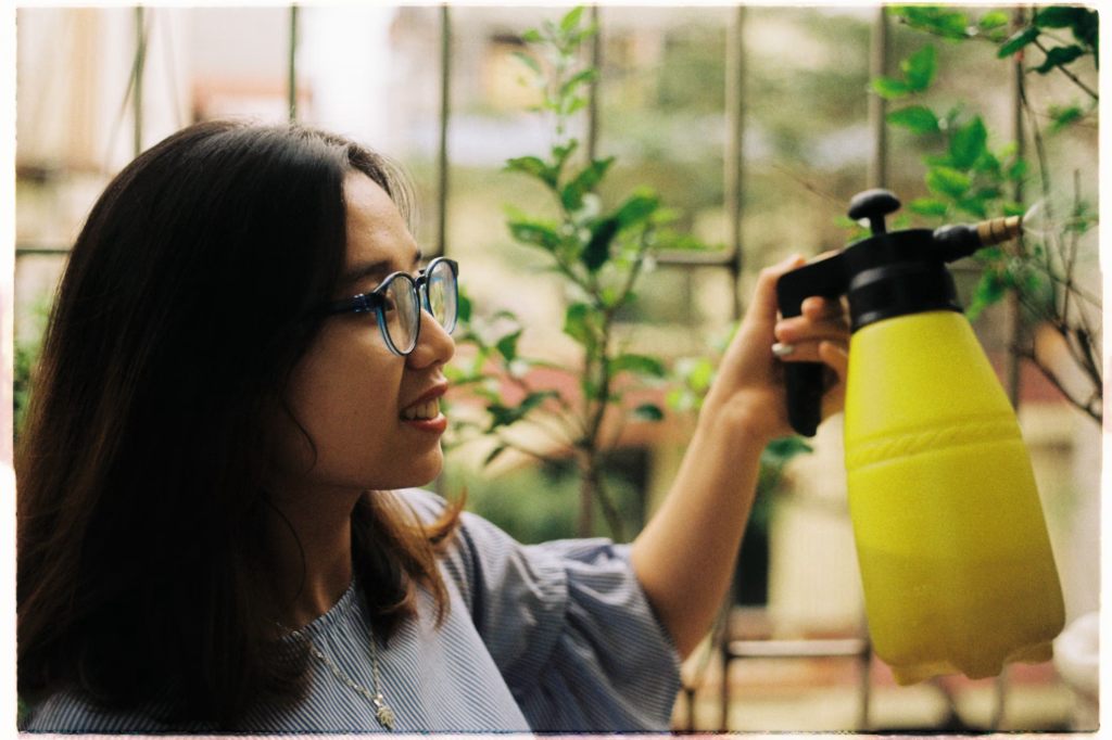 A woman with glasses is seen smiling as she sprays insecticides on a plant using a yellow spray bottle. The background features lush greenery and a blurred view of buildings, creating a serene and pleasant atmosphere. Her focused expression suggests her dedication to protecting the plants from pests.