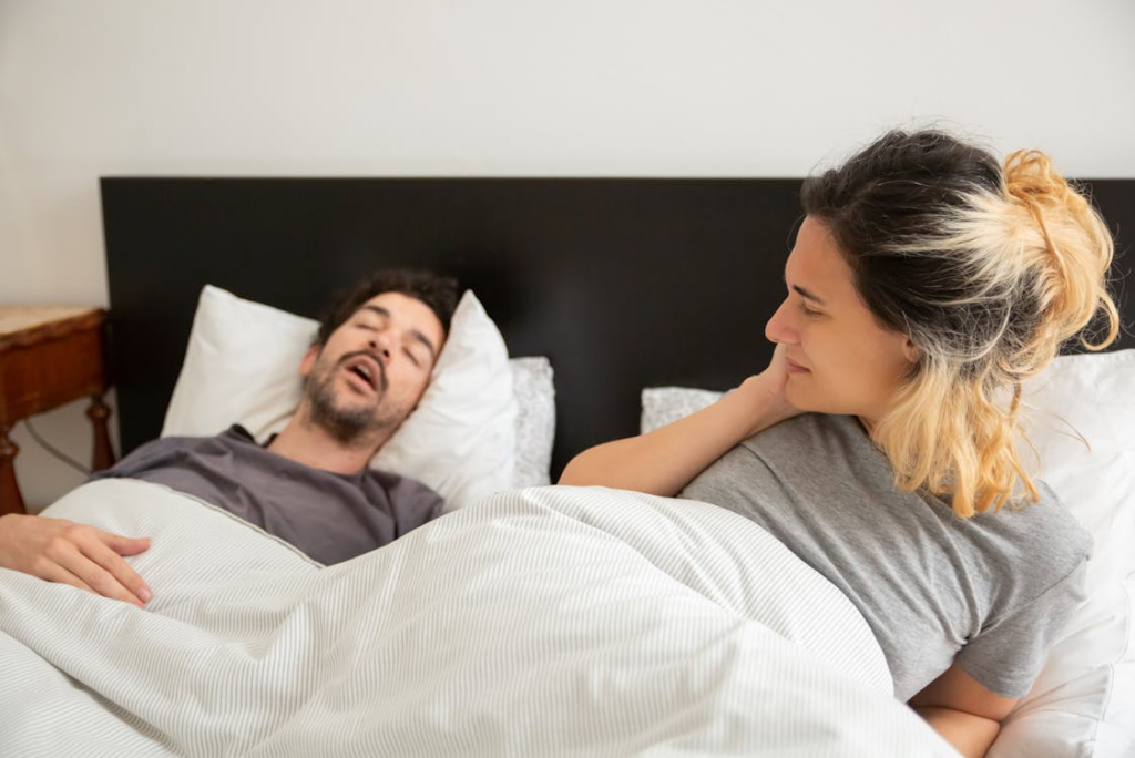 A woman lying awake in a well-lit bedroom, looking frustrated with her hand on her cheek, while a man sleeps beside her on white pillows, snoring.