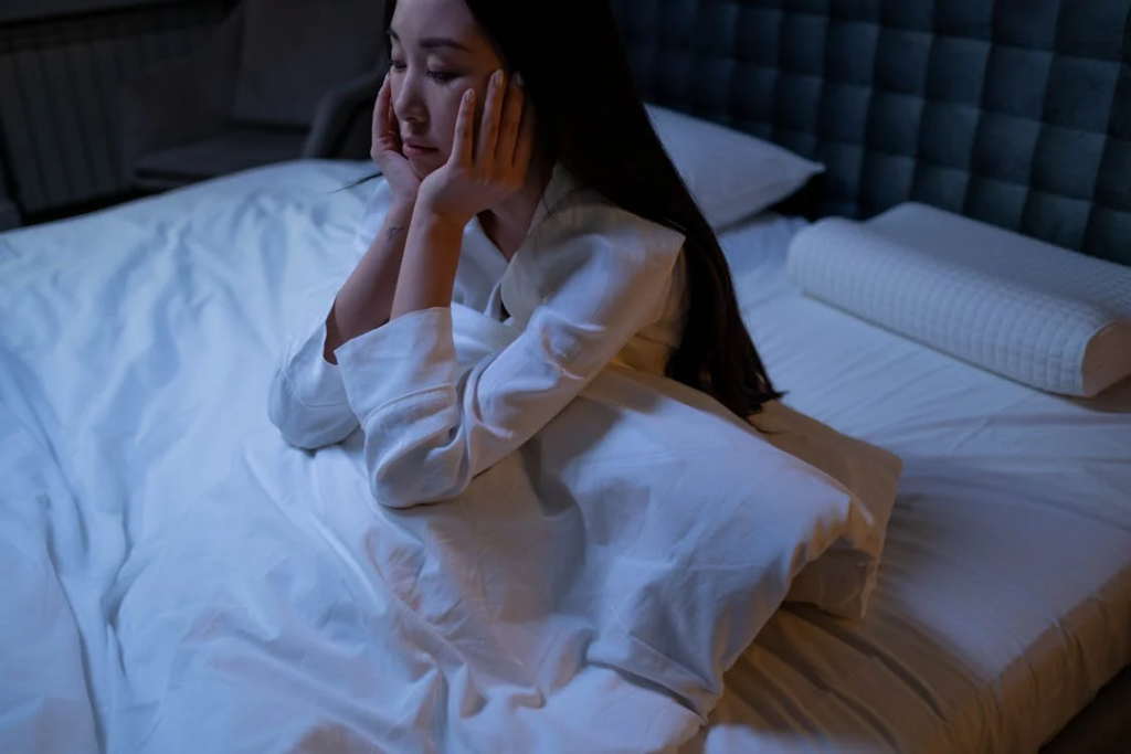 A woman sits in bed, appearing fatigued and distressed. Her hands are resting on her face, suggesting a sense of weariness or frustration. The dim lighting and disheveled bedding indicate that she may have had a restless night's sleep. This scene could represent common symptoms of sleep apnea, such as daytime tiredness, difficulty staying asleep, and a general feeling of exhaustion despite spending adequate time in bed.
