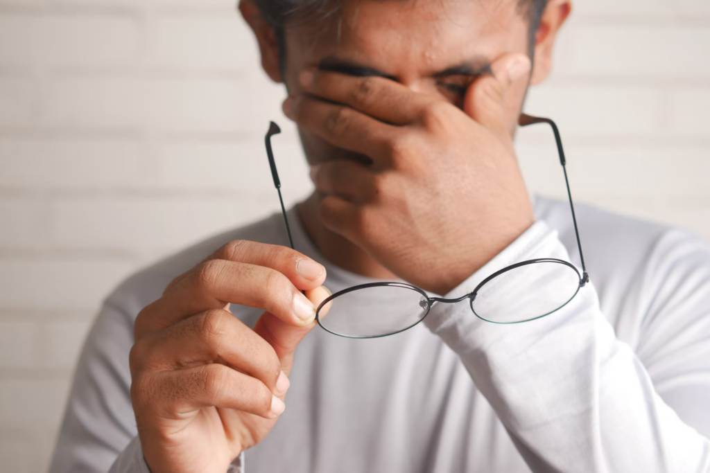 A man holding his glasses in one hand while rubbing his eyes with the other. He appears to be experiencing discomfort or fatigue, possibly due to eye strain or irritation. This could be indicative of symptoms related to prolonged screen time, dry eyes, or other vision-related issues. The setting is simple, with a plain background that keeps the focus on the man's actions and expression.