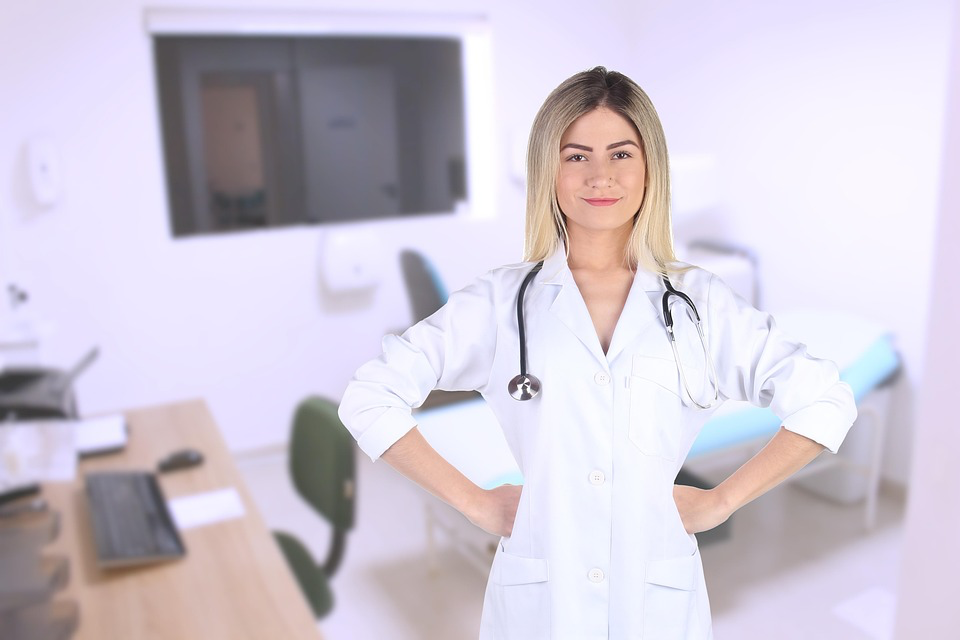 A medical professional standing confidently in a clinical setting. She is wearing a white lab coat, which is a common uniform for healthcare providers, and has a stethoscope around her neck, indicating her role in patient care. Her hands are placed on her hips, suggesting a sense of authority and readiness to assist. The background includes typical elements of a medical office, such as a desk with a computer, chairs, and examination equipment. 