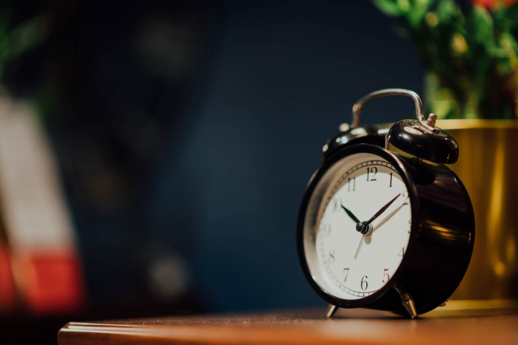 A classic black alarm clock with a white face displaying 10:10, sitting on a wooden surface. A golden planter or container is partially visible to the right, with some greenery blurred in the background. The scene has a warm, intimate atmosphere with a dark blue wall behind.