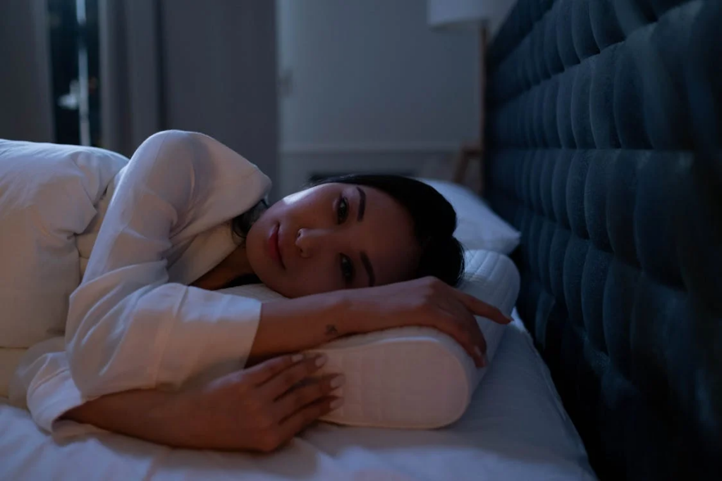 A woman lying awake in bed at night, looking restless with her eyes open. She's wearing white sleepwear and hugging her pillow while covered with a white comforter. A blue tufted headboard is visible in the background. This image illustrates poor sleep hygiene and insomnia struggles.