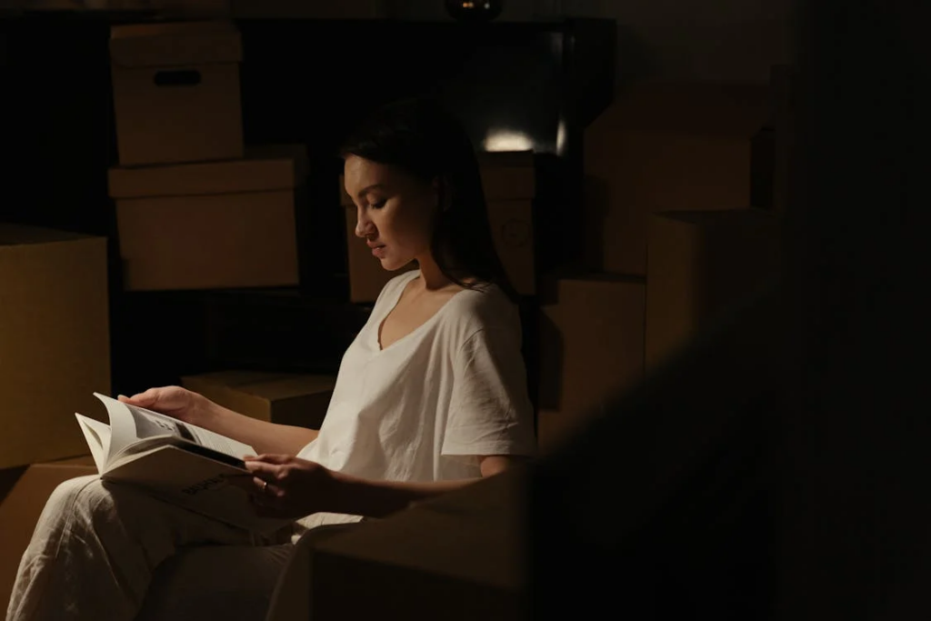 A woman with long dark hair in a white v-neck shirt sitting in low lighting, reading a book. She's surrounded by stacked cardboard moving boxes in what appears to be a dimly lit room. Her face is illuminated from the side as she focuses on the pages.