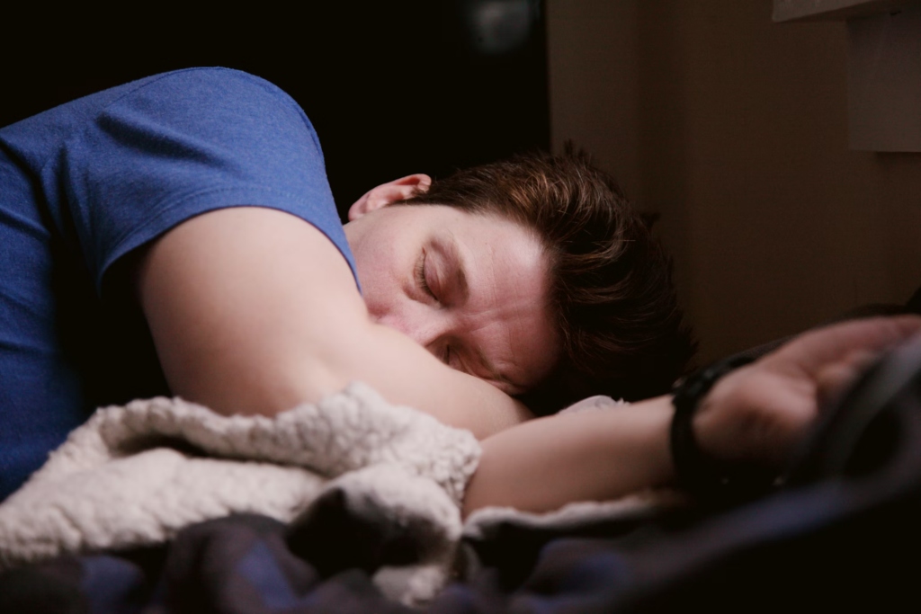 A close-up of a person with short brown hair peacefully sleeping. They're wearing a blue shirt and partially covered with a textured white blanket. The image captures deep sleep in a darkened room, demonstrating the restorative benefits of good sleep hygiene.
