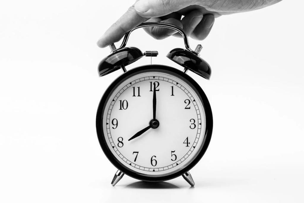 A black and white photograph of a hand holding a classic analog alarm clock showing 8:00. The clock has a white face with black numerals and traditional bell-style tops. The minimalist composition against a white background emphasizes the importance of consistent sleep schedules for proper sleep hygiene.