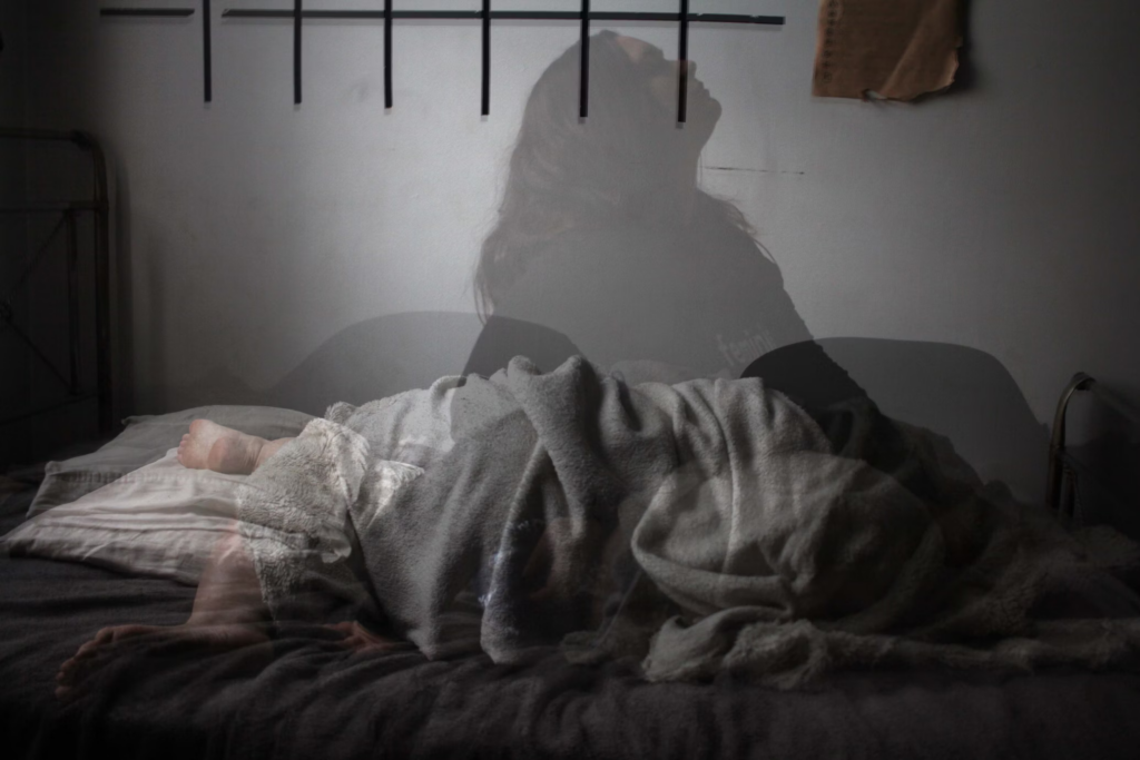 A haunting double-exposure image showing a person covered with a gray blanket on a bed, with their shadowy silhouette projected on the white wall above. Black horizontal bars are visible on the wall, creating a striking contrast. The image evokes feelings of sleep disturbance and restlessness.