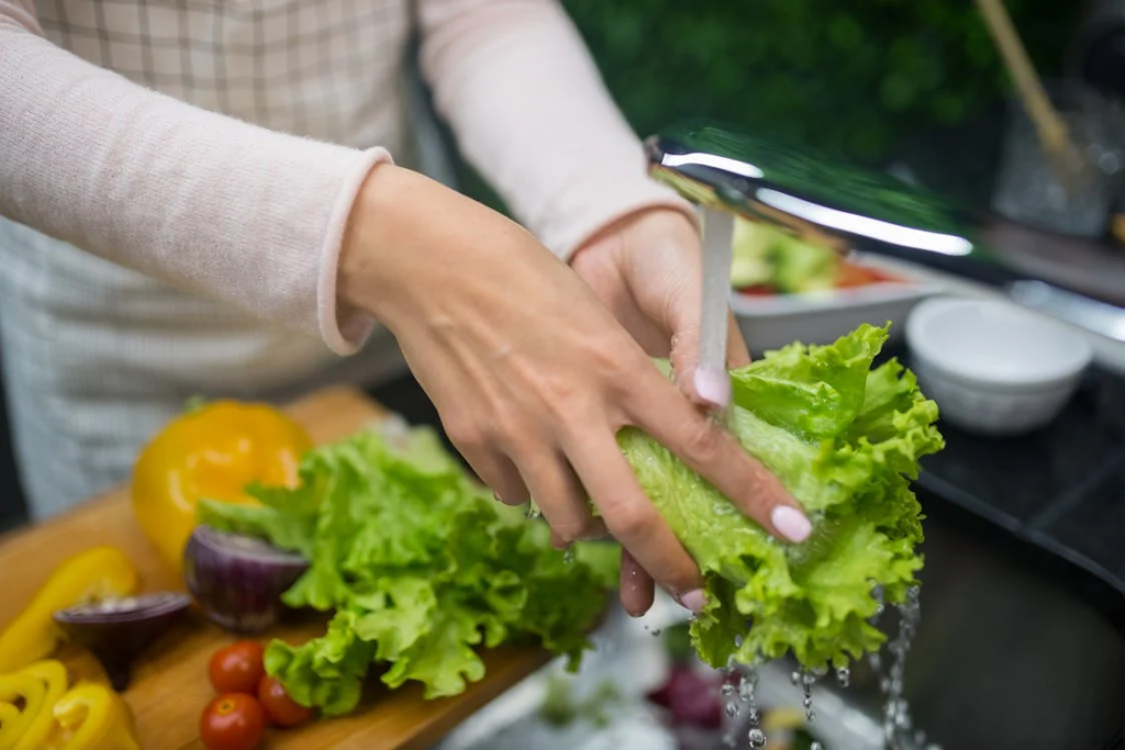 Close-up of hands washing fresh green lettuce leaves under running water. The person is wearing a light-colored long-sleeve top, and a wooden cutting board with various vegetables including yellow bell peppers, red onions, and cherry tomatoes is visible in the foreground. This image demonstrates proper food preparation and washing fresh produce.