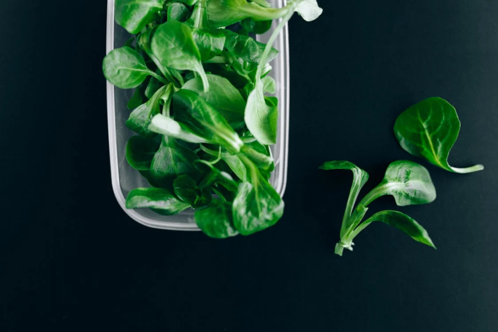 Fresh baby spinach leaves in a clear plastic container against a black background. Some individual spinach leaves are artfully arranged outside the container. The vibrant green color and tender leaves showcase the freshness of these pre-washed lettuce greens, ready for quick meal preparation or healthy salads.