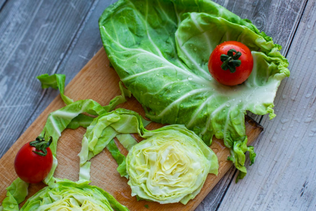 A wooden cutting board displaying fresh cabbage and lettuce ingredients. Two halved cabbage hearts with their layered pale green centers are positioned at the bottom, while a large green cabbage leaf with water droplets holds a bright red cherry tomato at the top. Another cherry tomato sits to the left against a weathered gray wooden background.