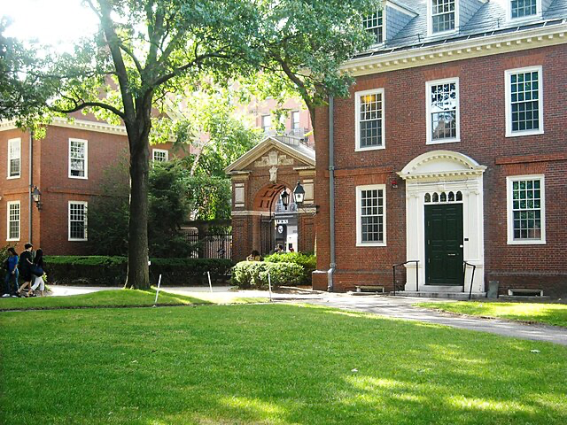 Historic red-brick Harvard University building with white-trimmed windows and doorway, surrounded by green lawn and shaded by trees.