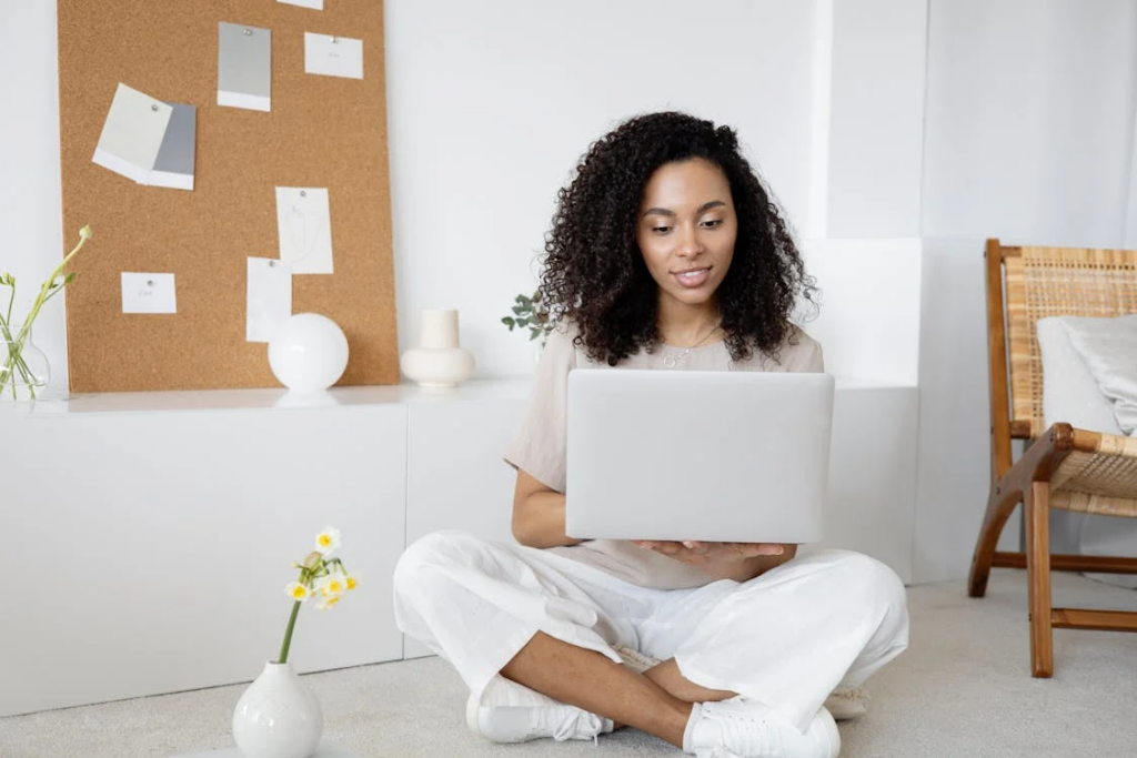 A young woman with curly hair sits cross-legged on the floor with a laptop, concentrating on her screen. She's wearing casual white pants and a light top in a clean, minimalist room with a cork board and small flower vase nearby.