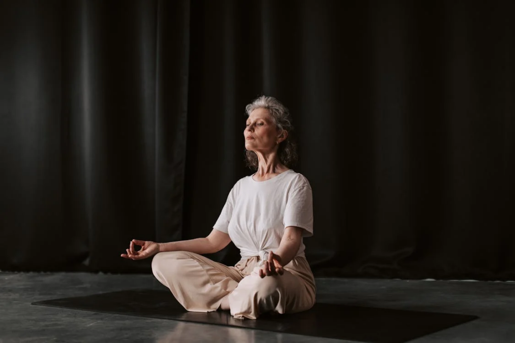Mature woman with silver hair in white clothing practicing meditation with Gyan Mudra hand positions. She sits cross-legged on a dark mat against a black backdrop, embodying serenity and mindfulness