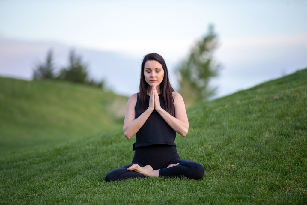 Woman in black attire meditating in prayer position on a grassy hillside at dusk. Her hands are pressed together in a prayer mudra as she sits in a cross-legged position against a backdrop of rolling hills.