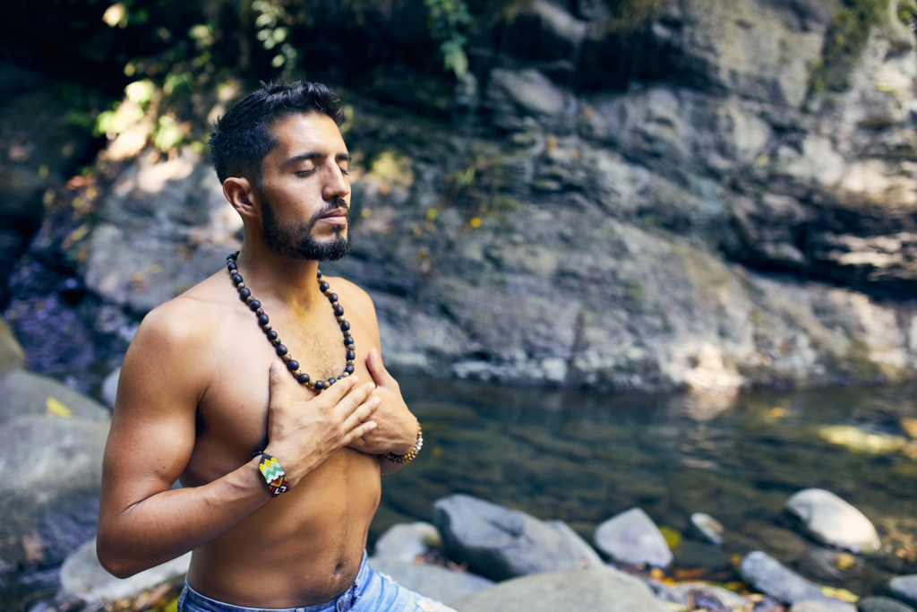 Man with beard meditating by a natural water pool with rocks, wearing a mala bead necklace. He stands shirtless with his hand on his heart in a spiritual gesture against a backdrop of stone formations.