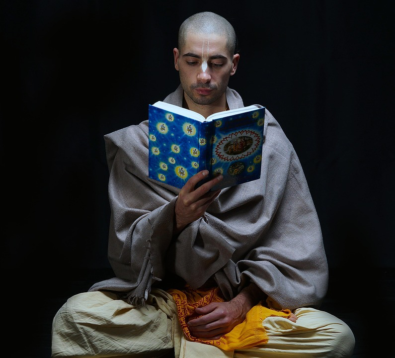Monk in light-colored robes reading a spiritual text with blue cover. He sits cross-legged in traditional meditation posture against a black background, fully absorbed in study.