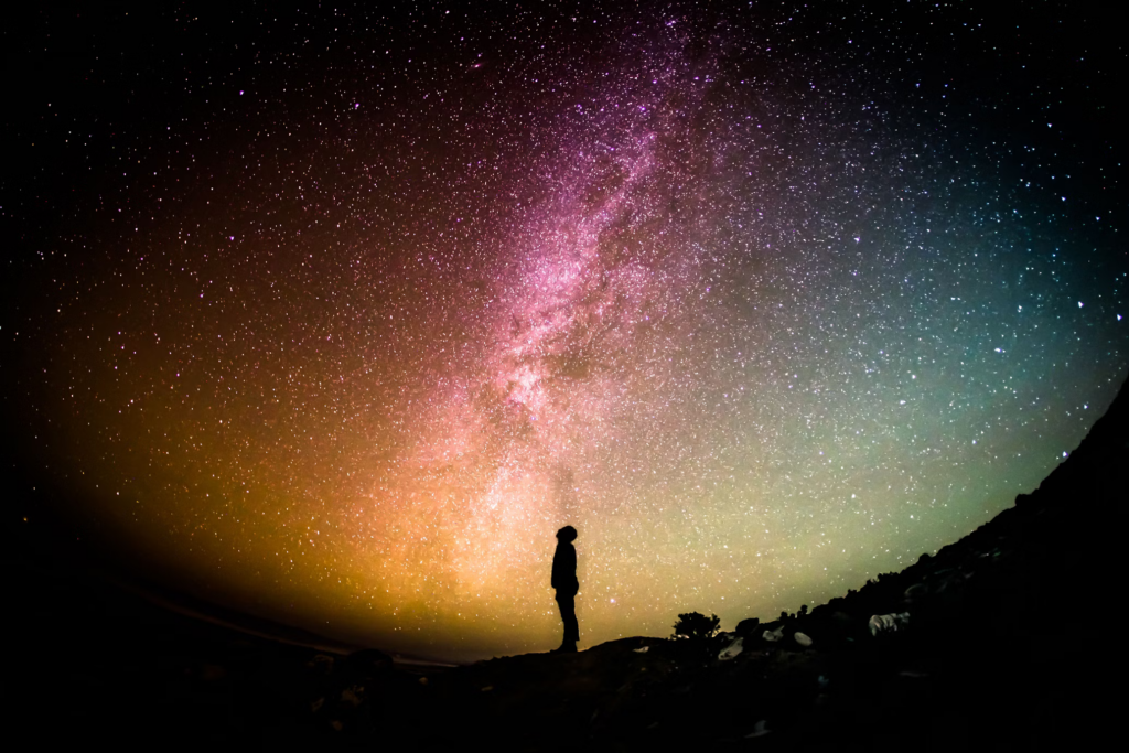 Silhouette of a person standing against a spectacular night sky filled with the Milky Way. Vibrant purple, orange and teal colors illuminate the cosmos, representing connection to universal consciousness.