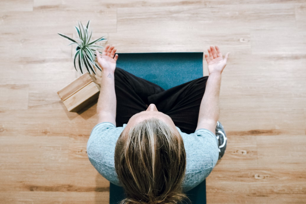 Overhead view of a person practicing yoga on a teal mat with a small plant nearby. The person is photographed from above showing the top of their head while seated in meditation.