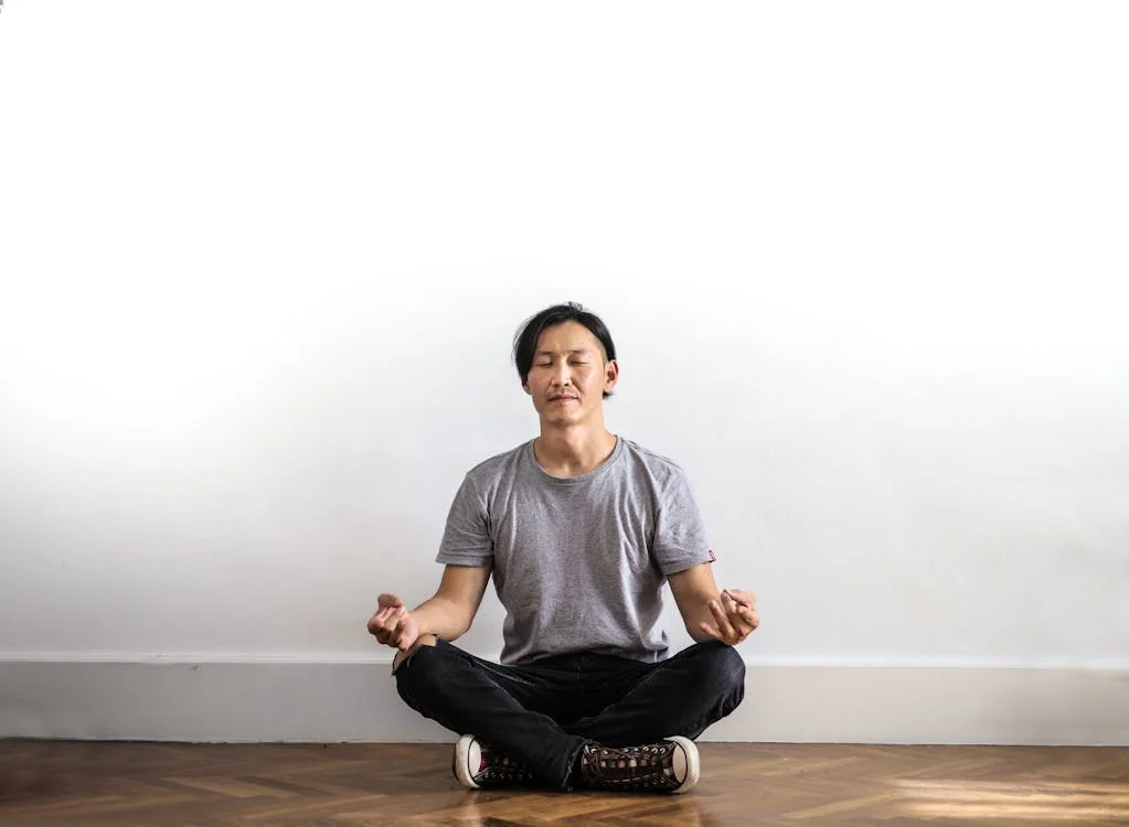 Man in gray t-shirt practicing Gyan Mudra meditation against a white wall. Sitting cross-legged on wooden flooring with eyes closed, he demonstrates the classic hand position where thumb and index finger form a circle.