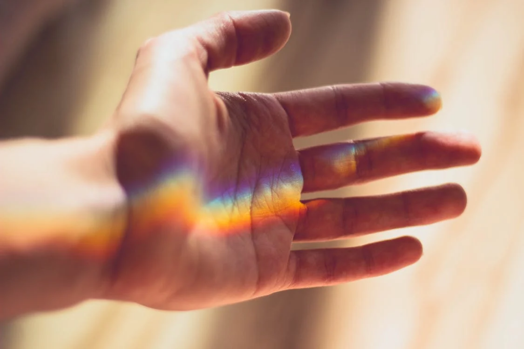 Close-up of a hand with a rainbow light prism reflecting across the palm. The spectrum of colors symbolizes the diverse energy elements connected to different fingers in Gyan Mudra practice.