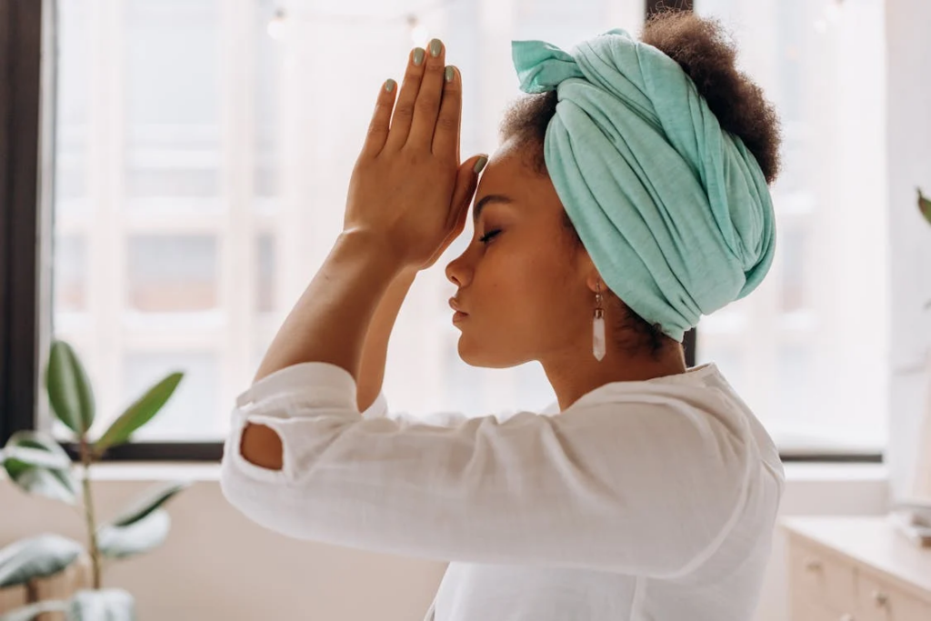 Woman with curly hair in turban practicing prayer mudra. Side profile shows her in white clothing with eyes closed and hands pressed together at forehead in mindful contemplation near a window.