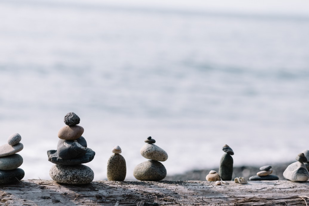 Balanced stone cairns on driftwood by the ocean. Several stacked rock formations of various sizes create a peaceful scene of balance and harmony against a blurred water background.