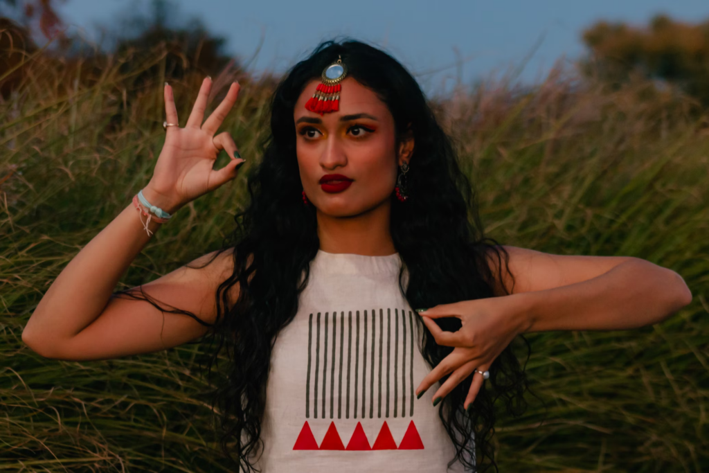 Woman with long dark hair displaying Gyan Mudra hand gesture in a natural setting at sunset. She wears traditional Indian-inspired clothing with geometric patterns and a decorative forehead ornament, demonstrating the ancient wisdom gesture with her right hand while standing among tall grasses.