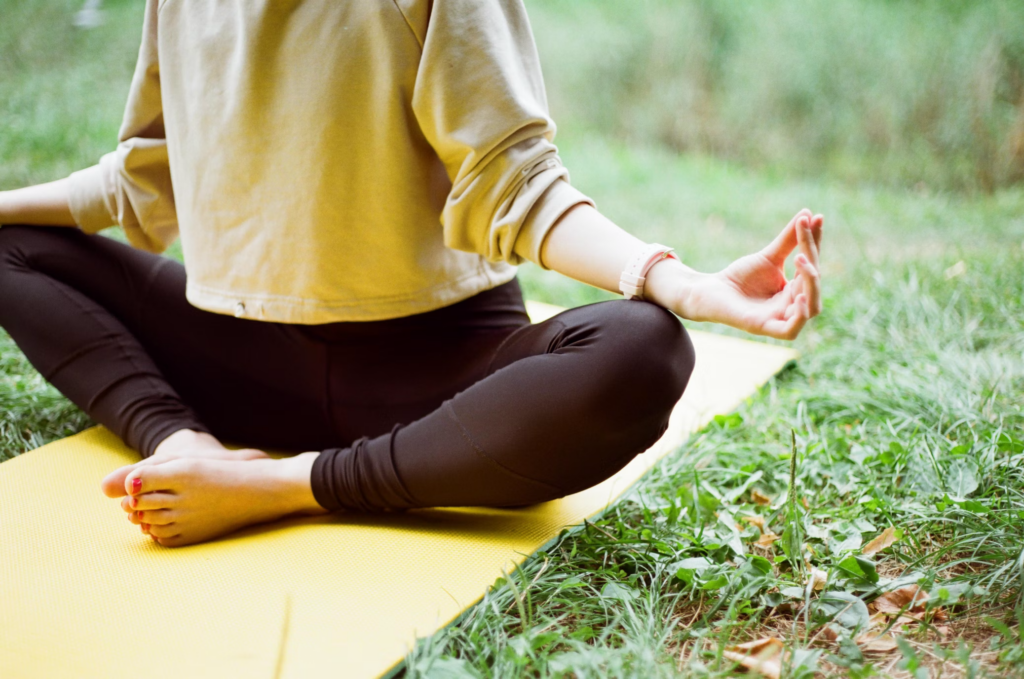 Person practicing Gyan Mudra meditation outdoors on a yellow yoga mat. Wearing a mustard-colored top and black leggings, they sit cross-legged on the grass with one hand forming the knowledge mudra where thumb and index finger connect in a circle, symbolizing the union of individual and universal consciousness.