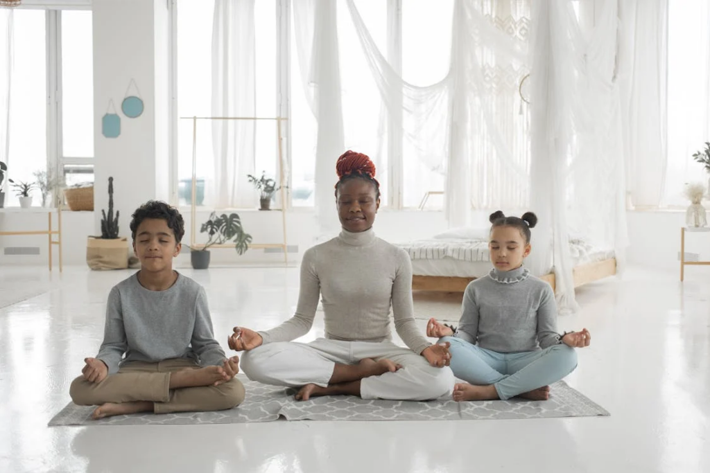 Family meditation scene in a bright, airy white room with three people practicing mindfulness together. A woman in white clothing sits between two children in gray tops, all in cross-legged positions with their hands resting in meditation mudras on their knees. Flowing curtains and plants create a serene atmosphere for their shared practice.