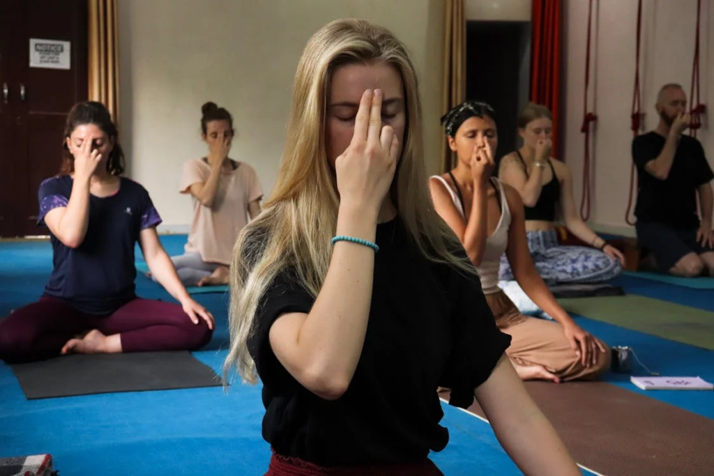 Yoga class practicing pranayama breathing techniques with participants in seated positions on colorful yoga mats. A woman with blonde hair in the foreground holds her fingers in a nostril-alternating breathing position, while other students behind her are shown in similar breath control exercises in a studio with blue flooring.