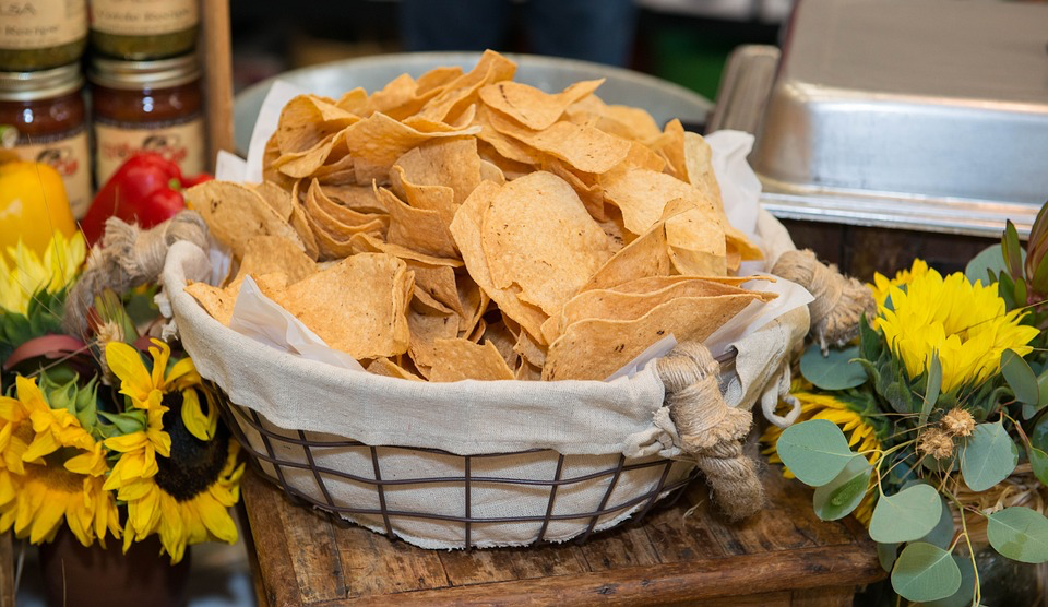 Golden tortilla chips piled high in a wire basket lined with white cloth, surrounded by bright yellow sunflowers.