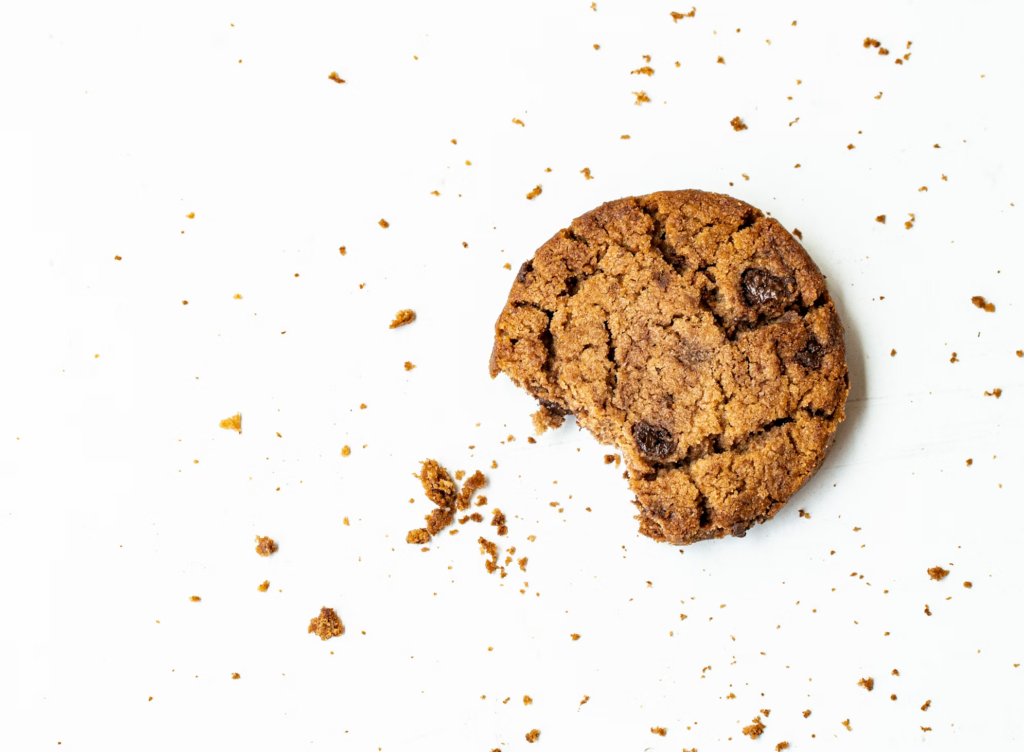 Partially eaten chocolate chip cookie with crumbs spread around it on a white background, showing a classic baked treat.