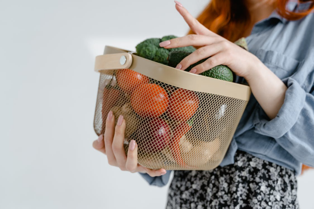 Mesh basket filled with fresh produce including green avocados and red tomatoes, held by hands wearing a blue shirt.